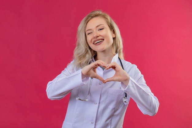 Smiling young doctor wearing stethoscope in medical gown showing heart gesture on red backgroung