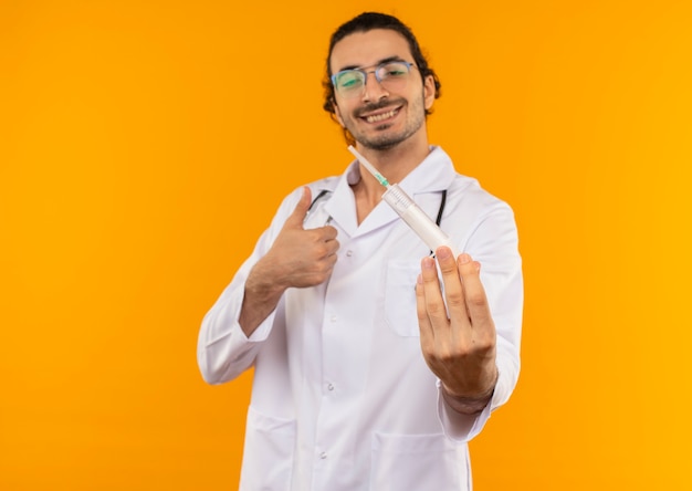 Free photo smiling young doctor with medical glasses wearing medical robe with stethoscope holding syringe his thumb up on isolated yellow wall with copy space