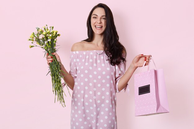 smiling young European woman has dark long wavy hair, wearing polka dot rose dress, holds gift bag and bouquet of flowers, posing on light pink, has birthday.