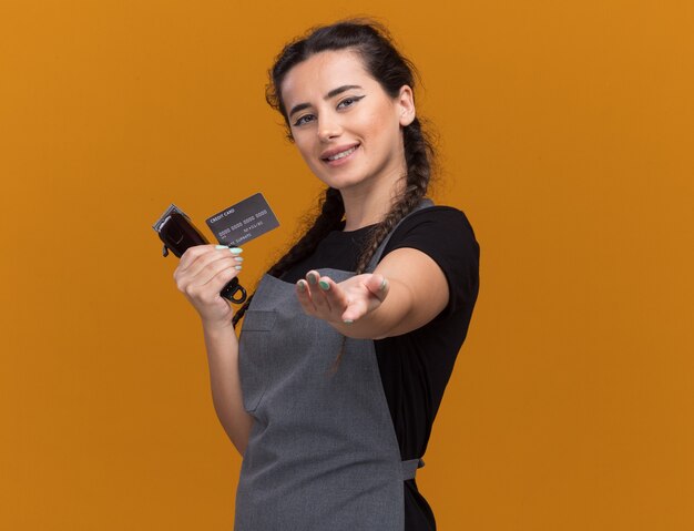 Smiling young female barber in uniform holding credit card and hair clippers holding out hand  isolated on orange wall with copy space