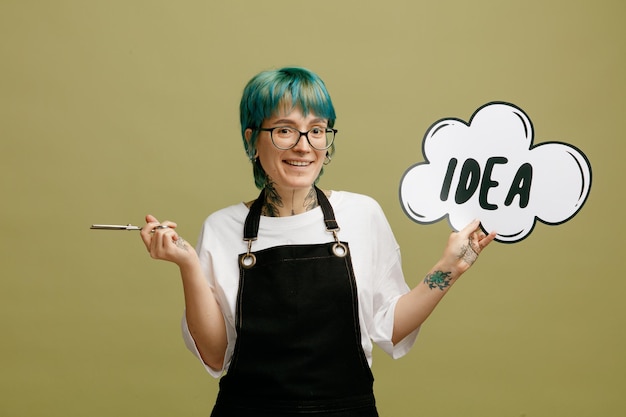 Smiling young female barber wearing glasses uniform showing idea bubble holding scissors looking at camera isolated on olive green background