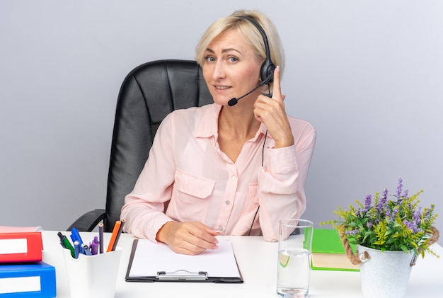 Smiling young female call center operator wearing headset sitting at table with office tools 