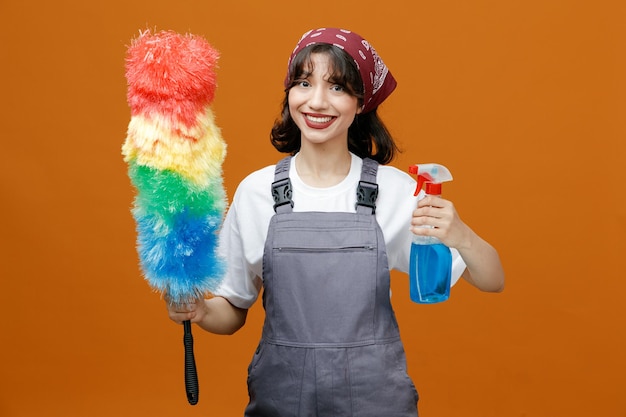 Smiling young female cleaner wearing uniform and bandana holding cleanser and feather duster looking at camera isolated on orange background