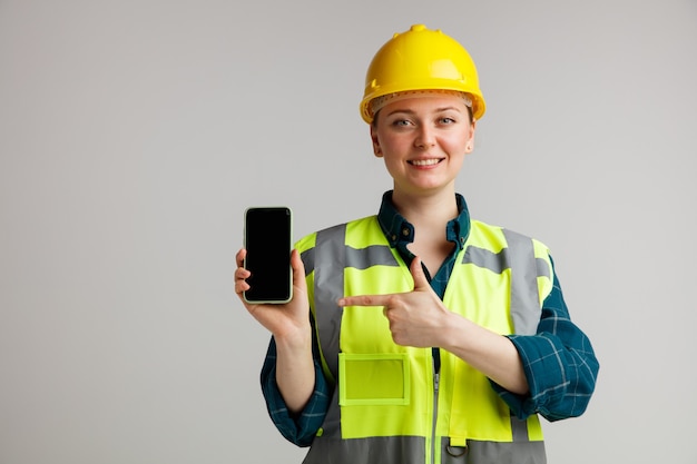 Smiling young female construction worker wearing safety helmet and safety vest holding and pointing at mobile phone 