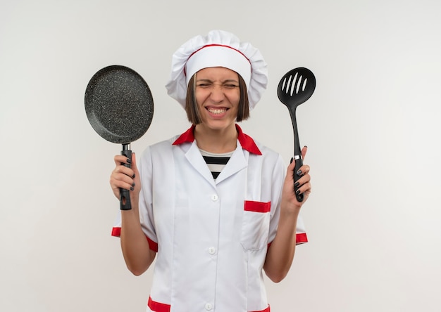 Smiling young female cook in chef uniform holding spatula and frying pan with closed eyes isolated on white wall