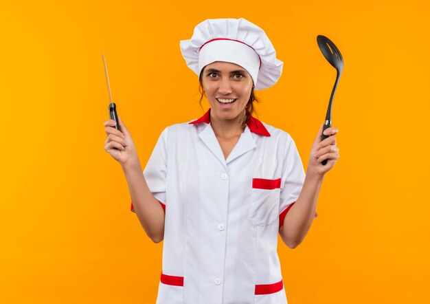 Smiling young female cook wearing chef uniform holding spatula and knife  with copy space