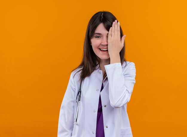 Smiling young female doctor in medical robe with stethoscope closes her eye with hand on isolated orange background with copy space