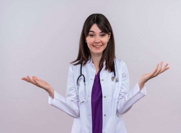 Smiling young female doctor in medical robe with stethoscope holds hands up on isolated white background with copy space