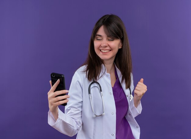 Smiling young female doctor in medical robe with stethoscope holds phone and keeps fist up on isolated violet background with copy space