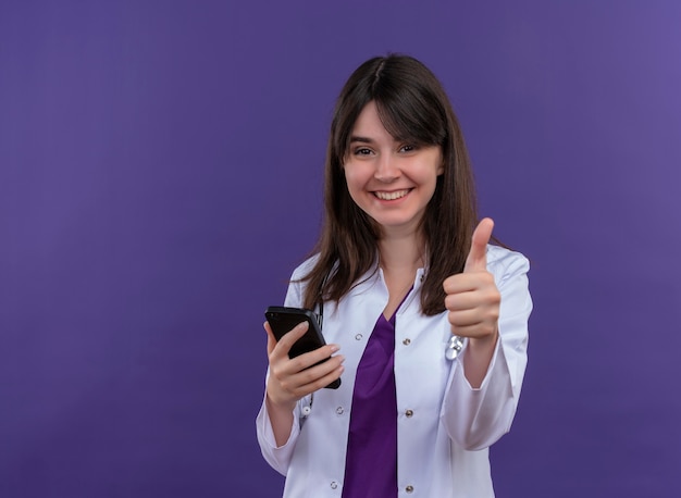 Smiling young female doctor in medical robe with stethoscope holds phone and thumbs up on isolated violet background with copy space