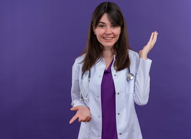 Smiling young female doctor in medical robe with stethoscope points down and raises hand up on isolated violet background with copy space