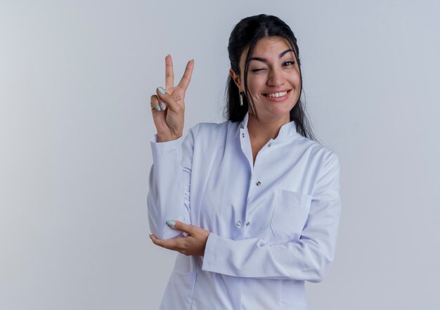 Smiling young female doctor wearing medical robe doing peace sign putting hand on elbow  isolated on white wall with copy space