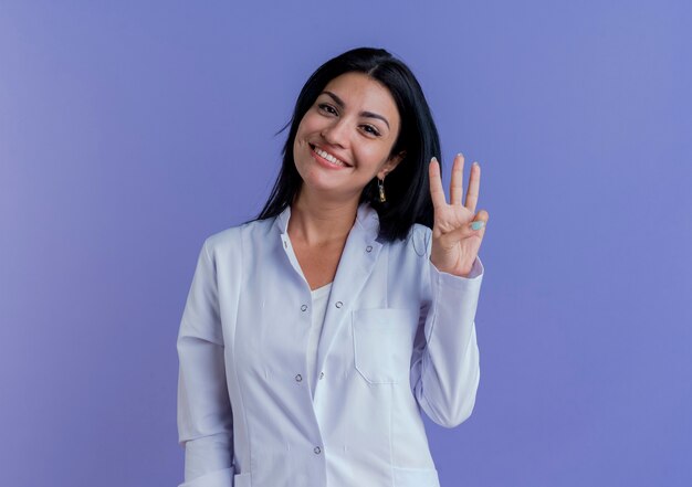 Smiling young female doctor wearing medical robe looking showing three with hand 