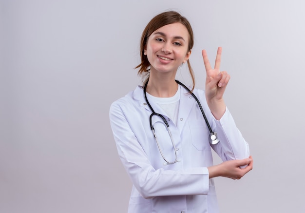 Smiling young female doctor wearing medical robe and stethoscope doing peace sign on isolated white wall with copy space