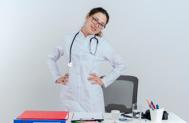 Smiling young female doctor wearing medical robe and stethoscope and glasses standing behind desk with medical tools looking keeping hands on waist isolated