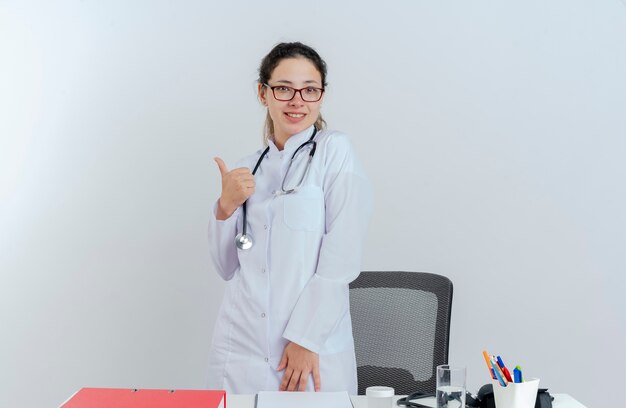 Smiling young female doctor wearing medical robe and stethoscope and glasses standing behind desk with medical tools looking showing thumb up isolated