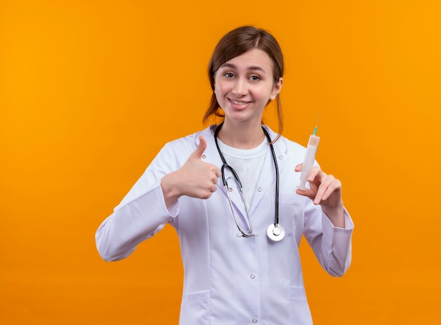 Smiling young female doctor wearing medical robe and stethoscope holding syringe and showing thumb up on isolated orange wall