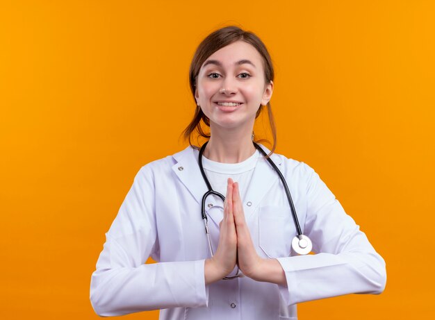 Smiling young female doctor wearing medical robe and stethoscope and putting hands together on isolated orange wall