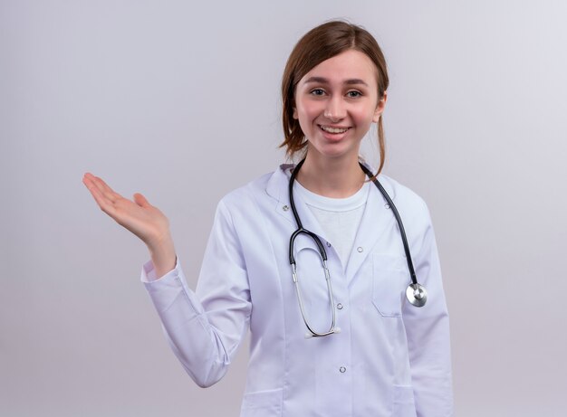 Smiling young female doctor wearing medical robe and stethoscope and showing empty hand on isolated white wall