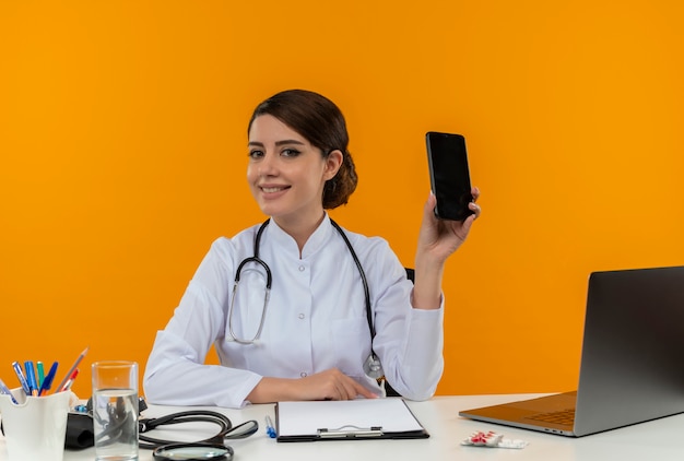 Smiling young female doctor wearing medical robe and stethoscope sitting at desk with medical tools and laptop showing mobile phone  isolated on yellow wall