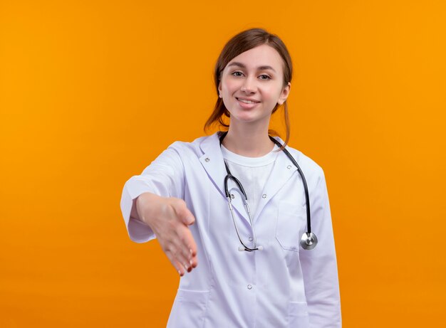 Smiling young female doctor wearing medical robe and stethoscope stretching hand  on isolated orange wall with copy space