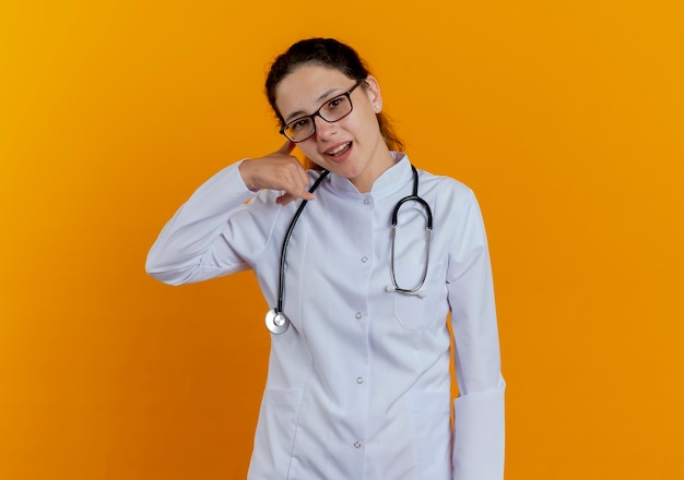 Free Photo smiling young female doctor wearing medical robe and stethoscope with glasses showing phone call gesture isolated on orange wall