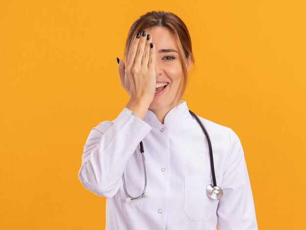 Smiling young female doctor wearing medical robe with stethoscope covered eye with hand isolated on yellow wall