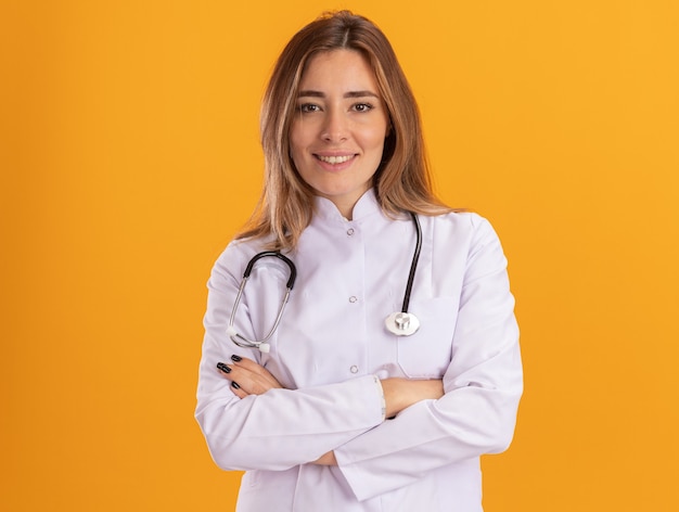 Smiling young female doctor wearing medical robe with stethoscope crossing hands isolated on yellow wall