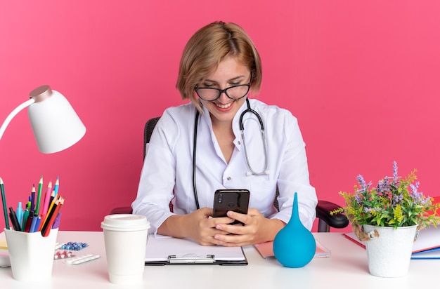 Free Photo smiling young female doctor wearing medical robe with stethoscope and glasses sits at desk with medical tools holding and looking at phone isolated on pink background