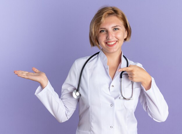 Smiling young female doctor wearing medical robe with stethoscope pretending holding and points at something isolated on blue background with copy space