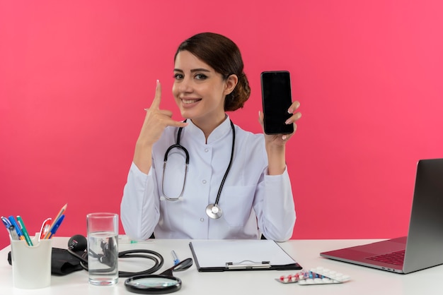 Free Photo smiling young female doctor wearing medical robe with stethoscope sitting at desk work on computer with medical tools holding phone and showing phone call gesture on pink wall