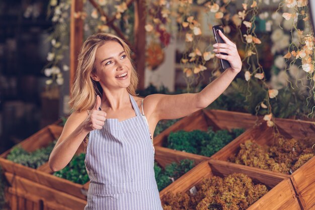 Smiling young female florist taking selfie on mobile phone showing thumb up sign in the shop