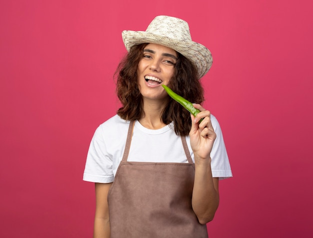 Smiling young female gardener in uniform wearing gardening hat holding pepper