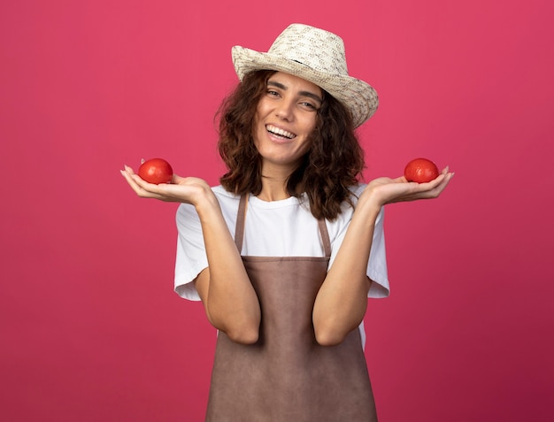 Free Photo smiling young female gardener in uniform wearing gardening hat holding tomatos