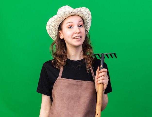 Smiling young female gardener wearing gardening hat holding rake 