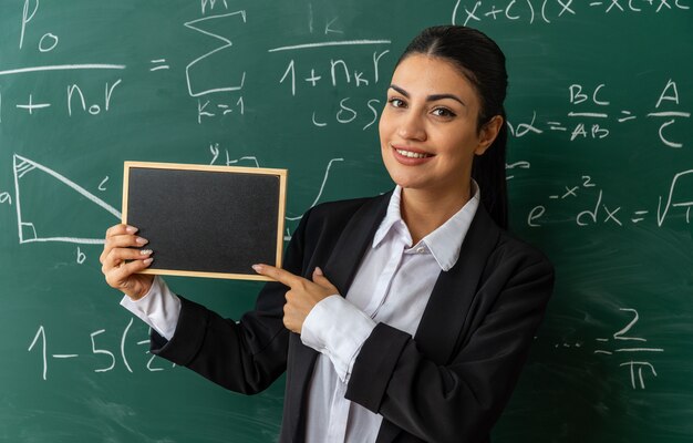 smiling young female teacher standing in front blackboard holding mini blackboard in classroom