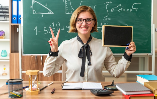 smiling young female teacher weaering glasses sits at table with school supplies holding mini blackboard showing peace gesture in classroom