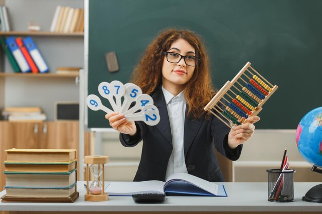 smiling young female teacher wearing glasses holding abacus with number fan sitting at desk with school tools in classroom