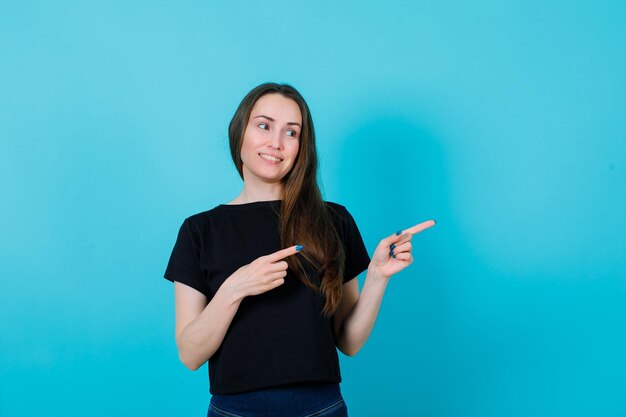 Smiling young girl is looking at right by pointing right with forefingers on blue background