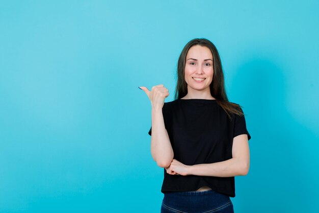 Smiling young girl is pointing left with thumb on blue background