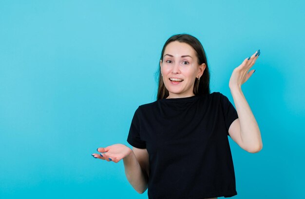 Smiling young girl is pointing up and down by opening wide hands on blue background