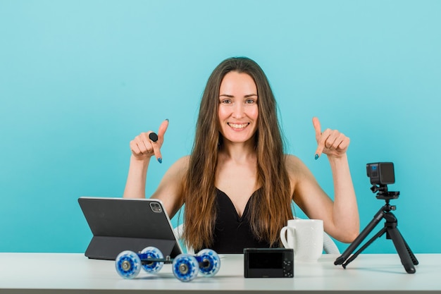 Smiling young girl is showing her camera and tablet with forefingers on blue background