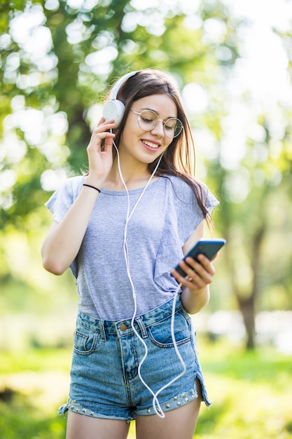 Smiling young girl student with backpack holding mobile phone, walking at the park, listening to music with earphones