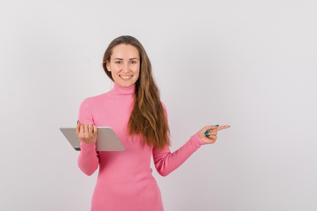 Smiling young girl with tablet computer is pointing to right on white background