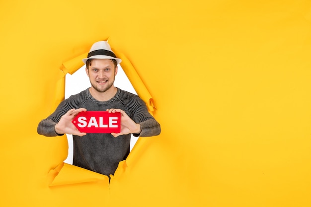 Free photo smiling young guy holding and showing sale sign in a torn on yellow wall