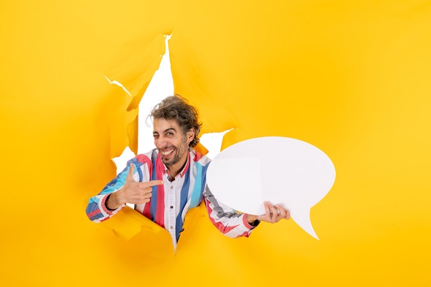 Smiling young guy pointing white page with free space and pointing something on the left side in a torn hole in yellow paper