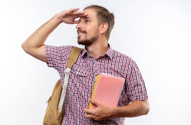 Smiling young guy student wearing backpack holding books looking at distance with hand isolated on white wall