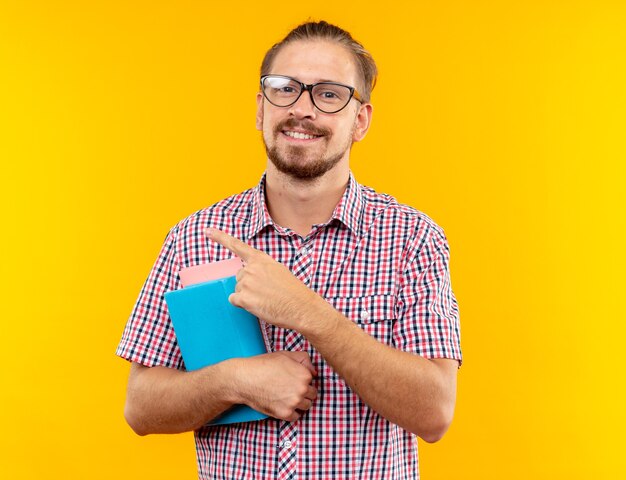 Smiling young guy student wearing backpack with glasses holding book points at side isolated on orange wall with copy space