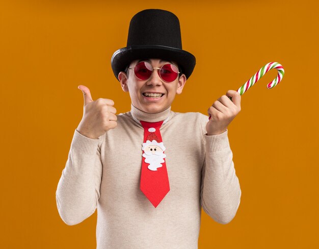 Smiling young guy wearing hat with christmas tie and glasses holding christmas candy showing thumb up isolated on yellow wall