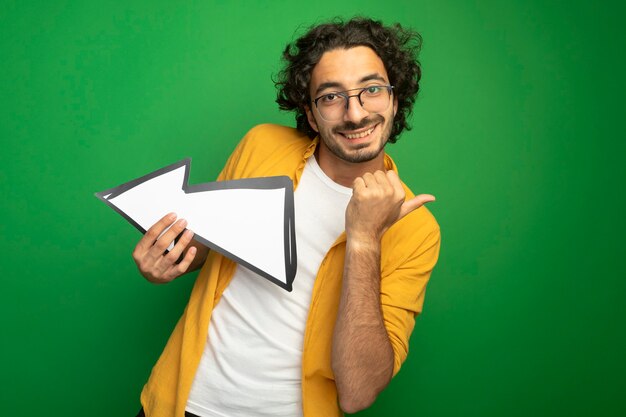 Smiling young handsome caucasian man wearing glasses holding arrow mark that is pointing at side looking at camera pointing at side isolated on green background with copy space